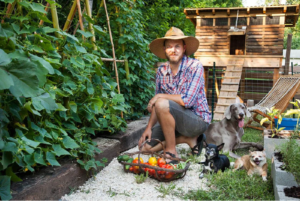 A man in a hat and shirt kneels in a lush garden, accompanied by dogs 